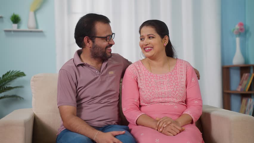 Happy smiling Indian middle aged couple posing or looking to camera at home on sofa - concept of married relationship, empty nesters and Quality Time.
