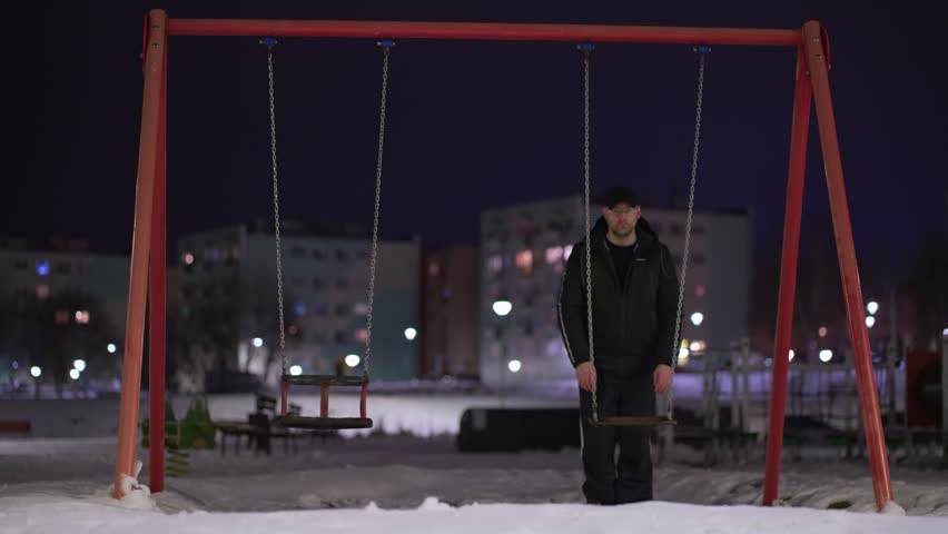 Mentally ill man with deep depression on children playground pushing swing on iron chains in cold winter night. Concept of child kids abduction