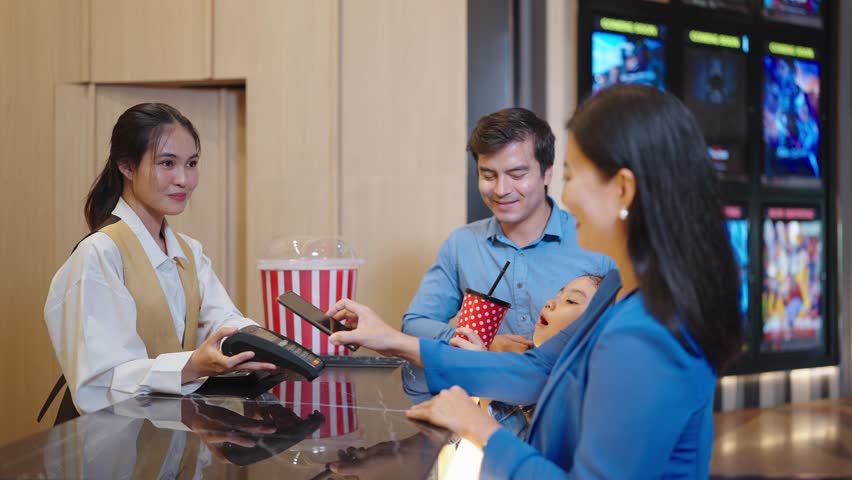 Happy Asian family choosing seats and buying movie ticket in cinema box office. Mother using smartphone scan for paying while buying movie ticket in cinema box office. Entertainment concept