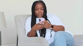 Carefree young african woman settled down in comfy wooden wicker armchair with cellphone enjoy weekend using internet e-shop web services, chat in social media network, spend free time alone at home