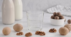 Vegan milk, pouring alternative milk made of walnuts into a glass on white wooden table, non-dairy product, kitchen table on background, 4k horizontal video, slow motion footage