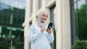 Young smiling muslim female employee in hijab is using a smartphone while walking on street near an office building. Happy female employee chatting online, texting with a friend, browsing social media