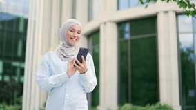 Young smiling muslim female employee in hijab is using a smartphone while walking on street near an office building. Happy female employee chatting online, texting with a friend, browsing social media - Powered by Shutterstock - Get 15% off with code: PIKWIZARD15