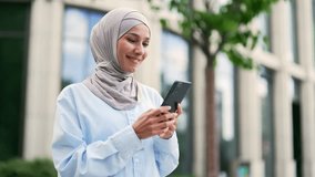 Close up. Young smiling muslim female employee in hijab is using a smartphone standing on the street near an office building. Happy woman chatting online, texting with colleague, browsing social media