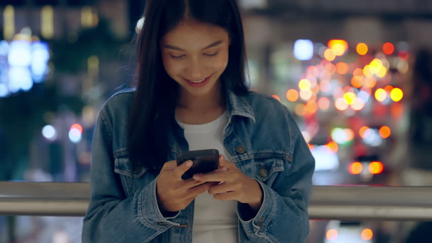 Beautiful young Asian woman standing in city street at night using mobile smartphone. Happy woman standing and typing messages on her mobile phone - Powered by Shutterstock - Get 15% off with code: PIKWIZARD15