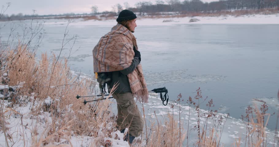 hicker holding Nordic walking poles watching flowing melting ice in the river in spring Slow motion Traveler standing resting on the bank of river adventure, guy feels cold, wrapped on tipped