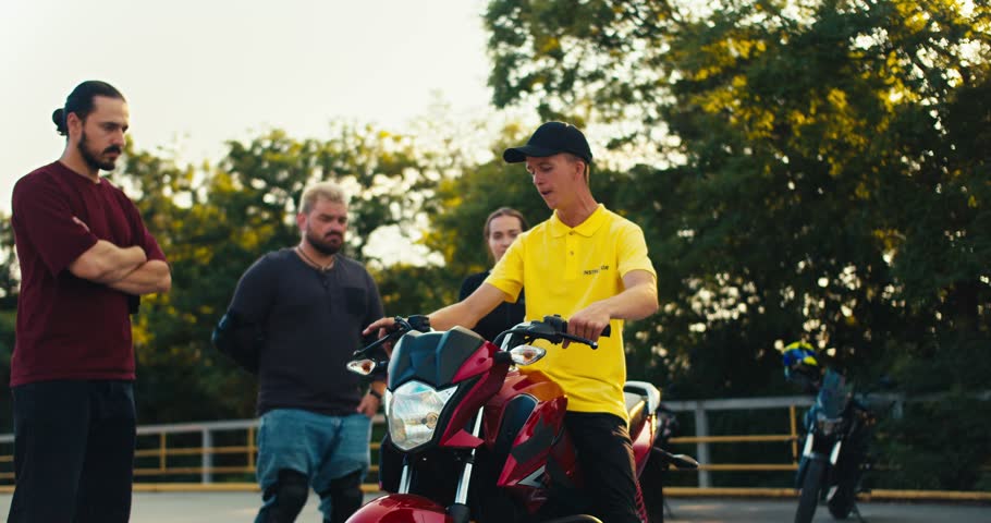 A driving instructor in a yellow T-shirt tells his students how to sit on a motorcycle and how to drive it correctly. Teachings at the site of a motorcycle school in sunny weather