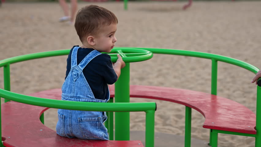 Adorable cute baby sitting on a merry go round. Little Caucasian child spending time on the playground in summer.