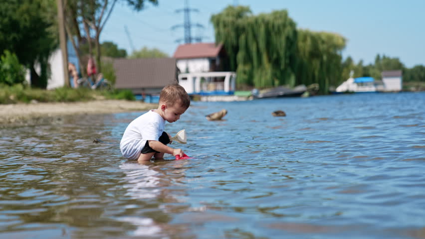 Little kid stands in river pouring water from his red watering can. Cute Caucasian toddler playing on the beach in summer.