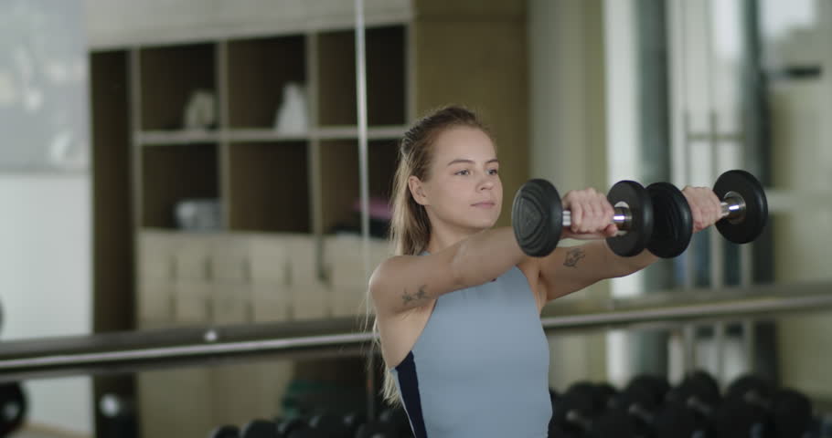 Fitness girl lifts weights in a sports club. A woman performs strength exercises with dumbbells in the gym, a beautiful fashionable blonde woman is engaged in fitness. Sports in the fitness room. 4K Royalty-Free Stock Footage #1108182785