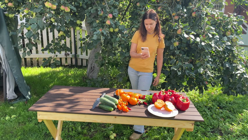 woman gardener taking smartphone photos of fresh vegetables on wooden table in summer garden