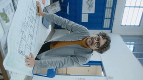 Male architect leaning on office desk with floor plan on it, looking at camera and smiling. Vertical clip, video portrait