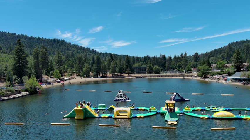 panning aerial footage of a gorgeous summer landscape with green lake water and kids playing on a jungle gym floating on the water, homes and green trees at Lake Gregory in Crestline California USA