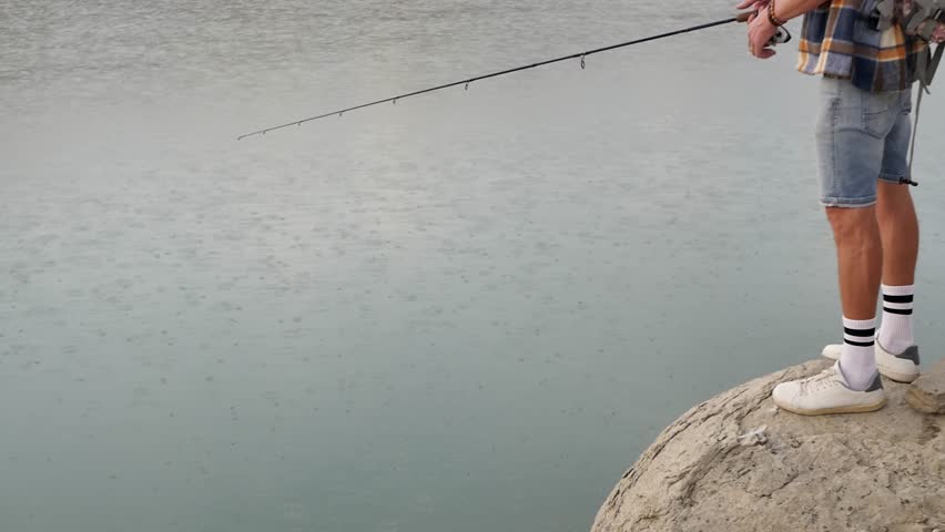 A man fisherman is fishing using a fishing rod while standing on a large stone on the shore of the lake and spinning the spinning reel. It rains while fishing. Young handsome guy fishing