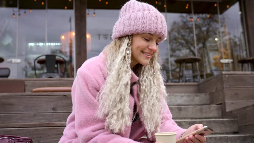 Fashionable mature woman in a pink knitted beanie hat and stylish coat outdoors in a cafe in autumn. Middle aged woman positive and smiling with pigtails in the city. Blur and selective focus