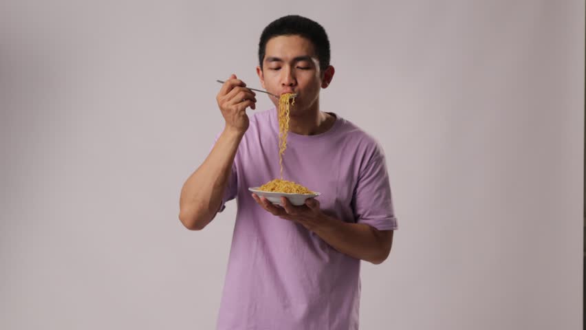 A man from Indonesia, Asia, wearing a purple t-shirt, appears to be enjoying noodles by slurping them. Isolated on a white background. Royalty-Free Stock Footage #1108220555
