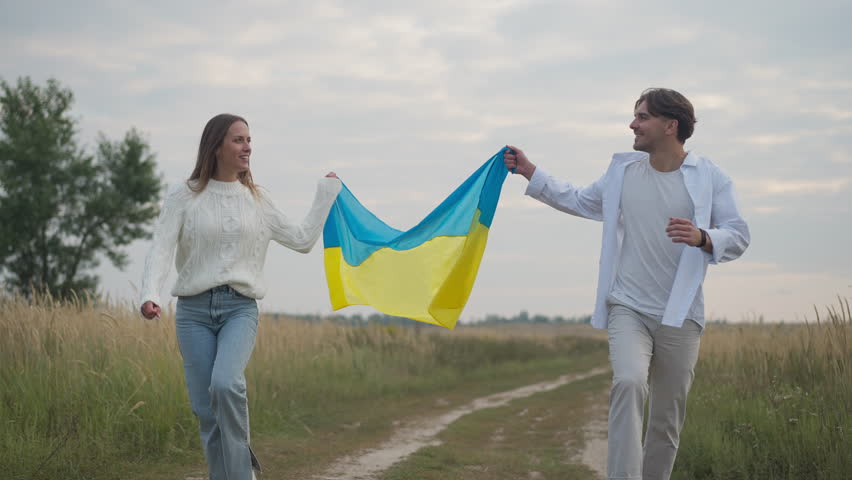Dolly shot joyful couple with Ukrainian flag running in slow motion in autumn field on overcast day. Front view portrait of smiling confident man and woman enjoying leisure having fun outdoors Royalty-Free Stock Footage #1108223207
