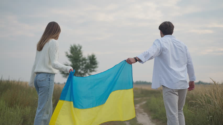 Tracking shot of joyful couple running in slow motion in field stretching Ukrainian flag. Wide shot back view happy cheerful young man and woman rejoicing outdoors Royalty-Free Stock Footage #1108223209