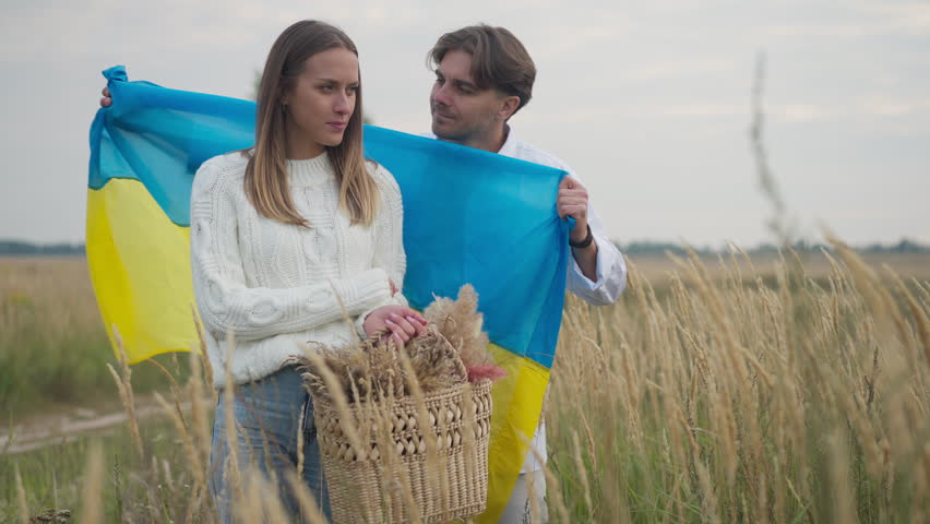Loving man covering woman with Ukrainian flag kissing cheek in slow motion. Portrait of beautiful Caucasian wife with wicker basket in wheat field with handsome husband Royalty-Free Stock Footage #1108223213