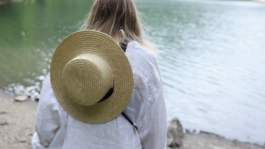 a girl with a straw hat on her shoulders and a white shirt walks by the lake and sits on a stone and looks at the lake