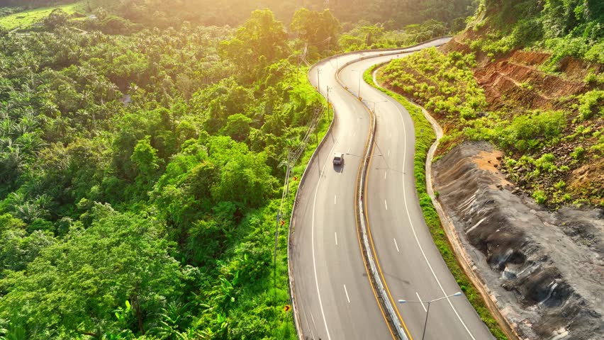 Experience the breathtaking allure of Ranong Province, Thailand, as a drone soars above a sunlit, winding asphalt road through lush tropical rainforests. The mesmerizing zigzag path. Nature concept.
 Royalty-Free Stock Footage #1108224539