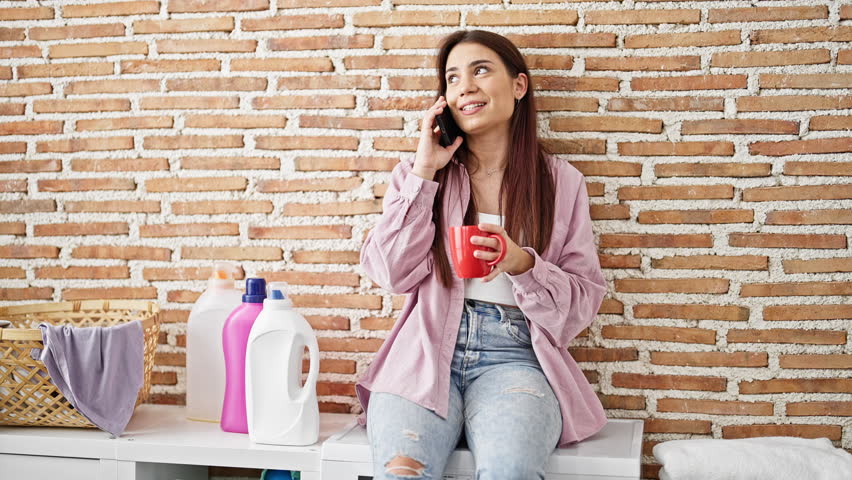 Young beautiful hispanic woman talking on smartphone drinking coffee waiting for washing machine at laundry room