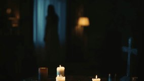 Video capturing a table with magical tools on it: candles, cross and beads. On the background there is a blurry female silhouette, ghost moving weirdly in front of the window.