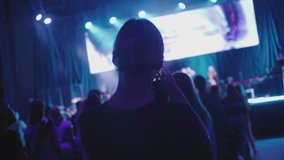 People at illuminated concert. A back view of a girl filming recording on a phone. Singing. Blurred stage. Worship christian concert with young people. Close up of mobile phone making a video of event