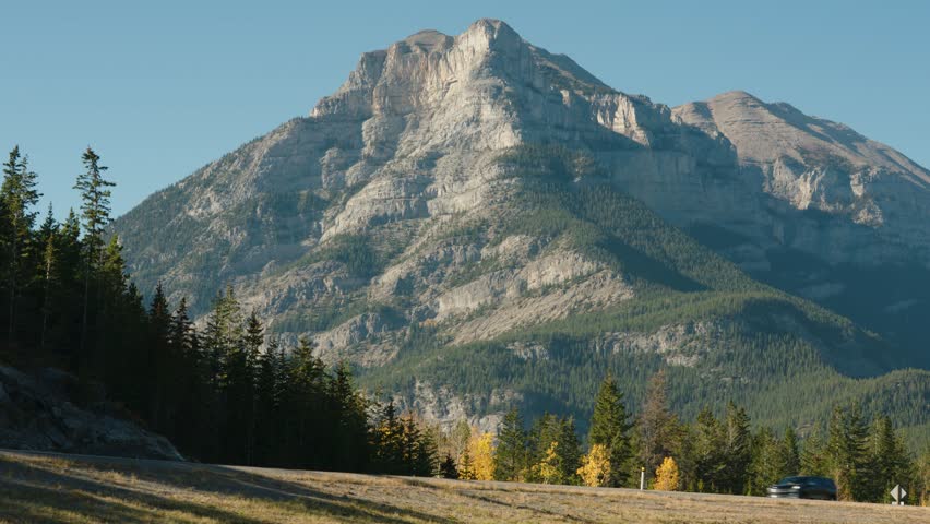 Cars Travel on a road in front of a large cliff face against a blue sky and forest near Canada