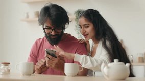 Arabian Indian couple woman girlfriend and man boyfriend talk using mobile phone make online order choose goods for apartment internet shopping delivery smiling multiracial family modern gadget addict