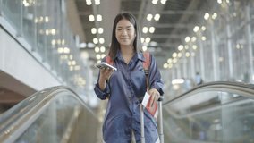 Young asian woman standing on escalator and making selfie using smartphone camera or video call at the airport, Smiling female talking in video conference by smartphone