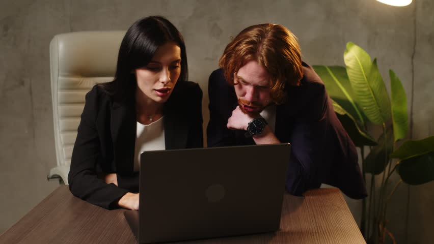 Portrait of a young woman and a business man sitting at a table late in the office in front of an open laptop and managing the company's finances and expenses. Royalty-Free Stock Footage #1108260677