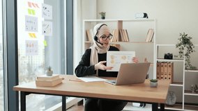 Focused arabian woman in headset pointing at document with infographics while having online presentation via laptop. Efficient manager in hijab showing sales report to colleagues in cozy workplace.