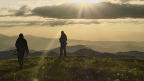 Silhouettes of men on a mountain at sunset. The wind ruffles the grass. The sun is breaking through clouds. Silhouettes of mountain peaks in background. Epic video of mountain tourism. Slow motion