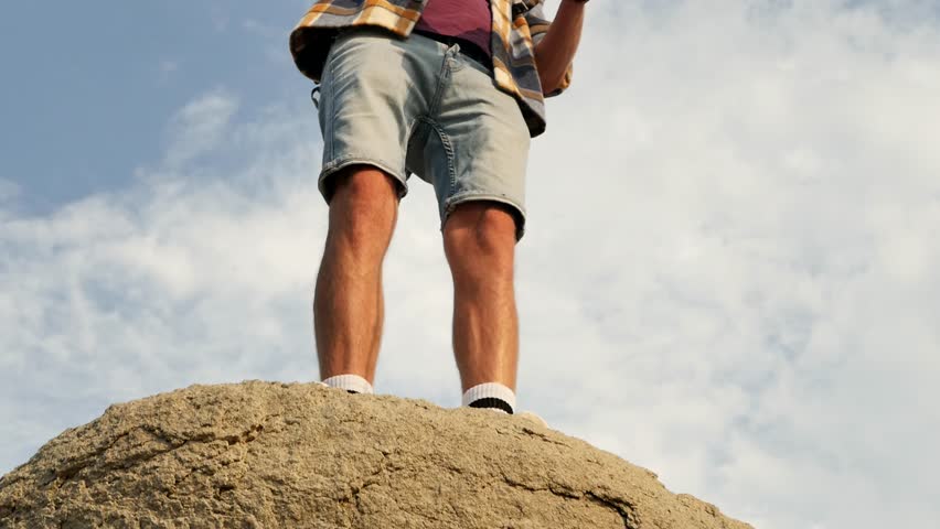 Looking up, the camera pans up to capture a fisherman standing on a large rock against the sky and reeling in his line while fishing for predatory fish in fresh water. Young handsome stylish man