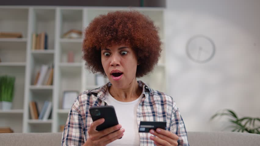 Overjoyed african american woman holding credit card and modern smartphone while getting great news. Surprised brunette sitting on couch and gesturing from happiness during win lottery notification Royalty-Free Stock Footage #1108263899