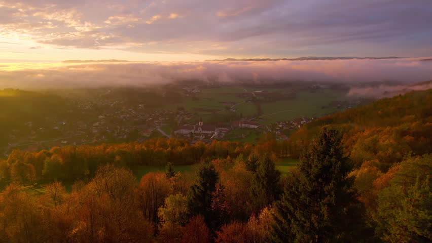 AERIAL: Majestic morning over village with rolling fog further in the valley. Beautifully colored autumn forest trees glowing in golden sunlight on a hill above vale in an idyllic countryside setting. Royalty-Free Stock Footage #1108264039