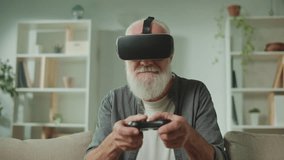 An Old Man in VR Glasses Plays Computer Games With a Joystick, Sitting in the Sofa with a Popcorn.A Modern Elderly Man Plays PlayStation. Augmented Reality and Gaming for Seniors.