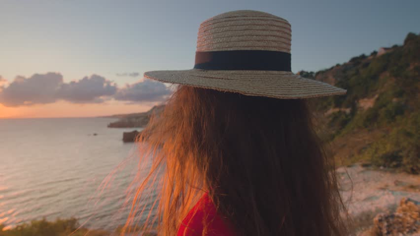 An attractive young Caucasian woman with long hair in a red dress and a straw hat looks at the sun setting in the sea. The wind blows her hair. Realtime