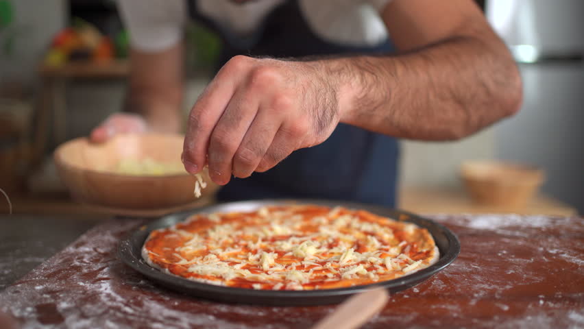Happy cheerful caucasian man making - baking a pizza at home, man adding an ingredients over the pizza round in a tray before put it in the oven.