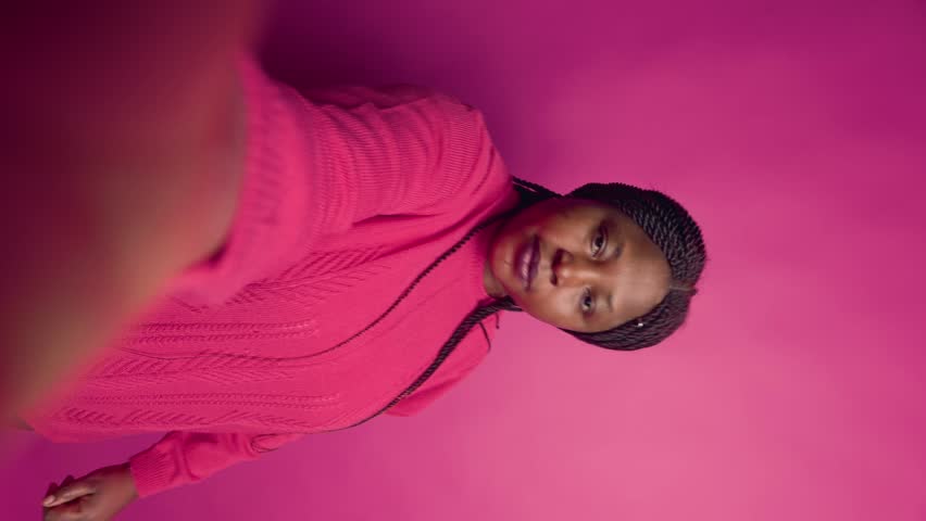 vertical video African American fashion blogger doing adorable hand motions on video chat. Pretty black woman taking selfies with mobile device while wearing pink jumper against isolated backdrop. Royalty-Free Stock Footage #1108272983
