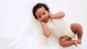 a close-up portrait of a small African-American baby girl in a white bodysuit on a cotton bed at home, a funny six-month-old smiling joyful black newborn baby lies on the back - Powered by Shutterstock - Get 15% off with code: PIKWIZARD15