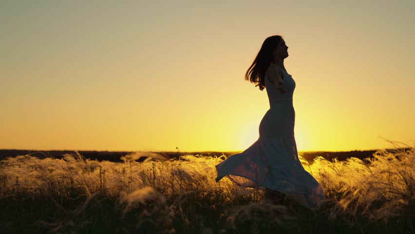 Happy young woman joyfully dances spinning with her arms outstretched at sunset. Concept of womens dreams success travel, flight. Beautiful girl, holding her hands to sides, dances on field under sun
