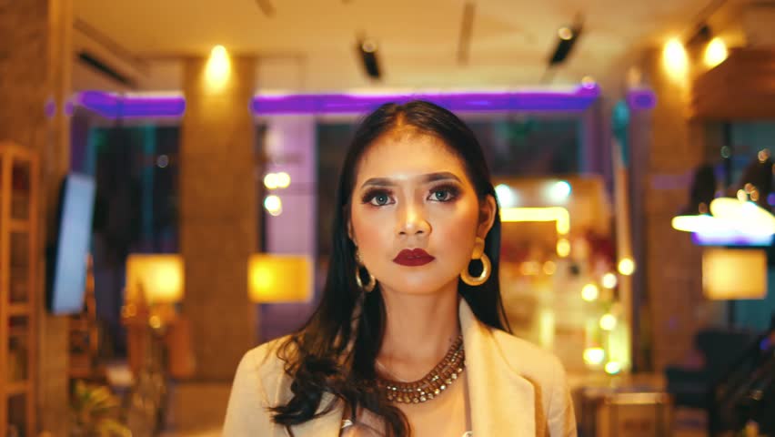 Tracking shot following a glamorous Asian woman walking towards the camera through a brightly lit, luxurious hotel lobby at night, looking around confidently.