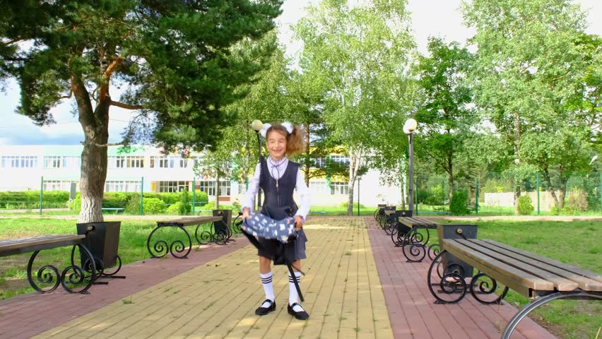 Cheerful funny girl with toothless smile in school uniform with white bows in school yard tosses the backpack. Back to school, September 1. Happy pupil. Primary education, elementary class.

