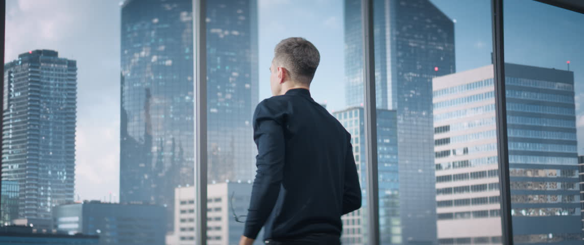 Caucasian Businessman Sitting Down At His Desk And Starts Typing On Laptop Computer In Corner Office With Megapolis Cityview. Proffesional Digital Entreprenuer Working With Ecommerce. Anamorphic Shot