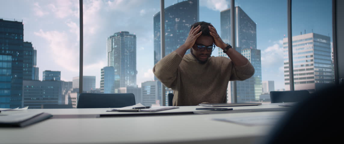 Stressed Out Balck Businessman at a Desk in Modern Skyscraper Office, Using Laptop Computer. Tired Manager Completing Complicated Financial Report Before Deadline, Feeling Overwhelmed. Anamorphic Shot
