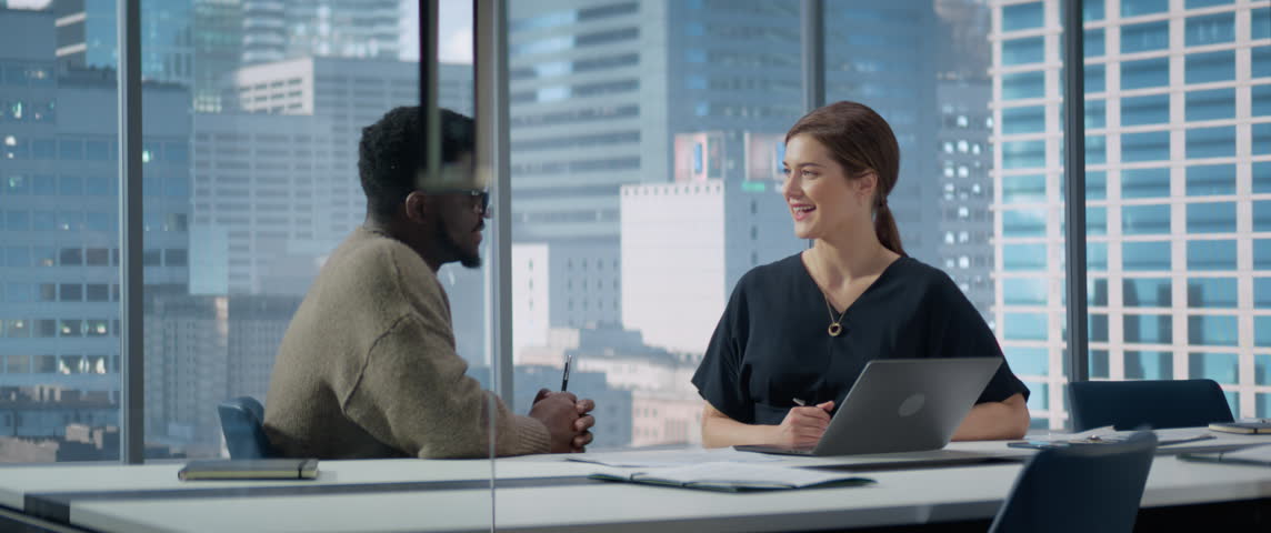 Businesspeople in Skyscraper Office: Business Meeting of Two Managers Using Laptop. Female CEO and African American Operations Director Discuss Objectives or Financial Plans. Anamorphic Shot
