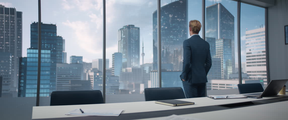 Caucasian CEO Entering His Corner Office With Big City View. Male Entrepreneur Standing And Looking Out Of Window Thoughtfully, Planning Business Decisions, Coming Up With Ideas. Anamorphic Shot