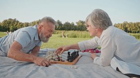 Side view of elder husband and wife enjoying active retirement life and spending time on nature. Casually dressed senior couple lying on street rug and competing in game of chess. - Powered by Shutterstock - Get 15% off with code: PIKWIZARD15